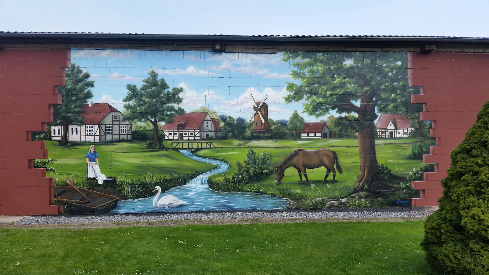 Fassaden Gestaltung Wendland. Ein großes Wandbild mit einer Landschaft. Im Hintergrund ein Dorf mit Fachwerkhäusern und einer Mühle. Im Vordergrund ein Bach mit einem Schwan, ein Pferd grast auf deiner Wiese und ein Ruderboot liegt am unteren linken Rand.